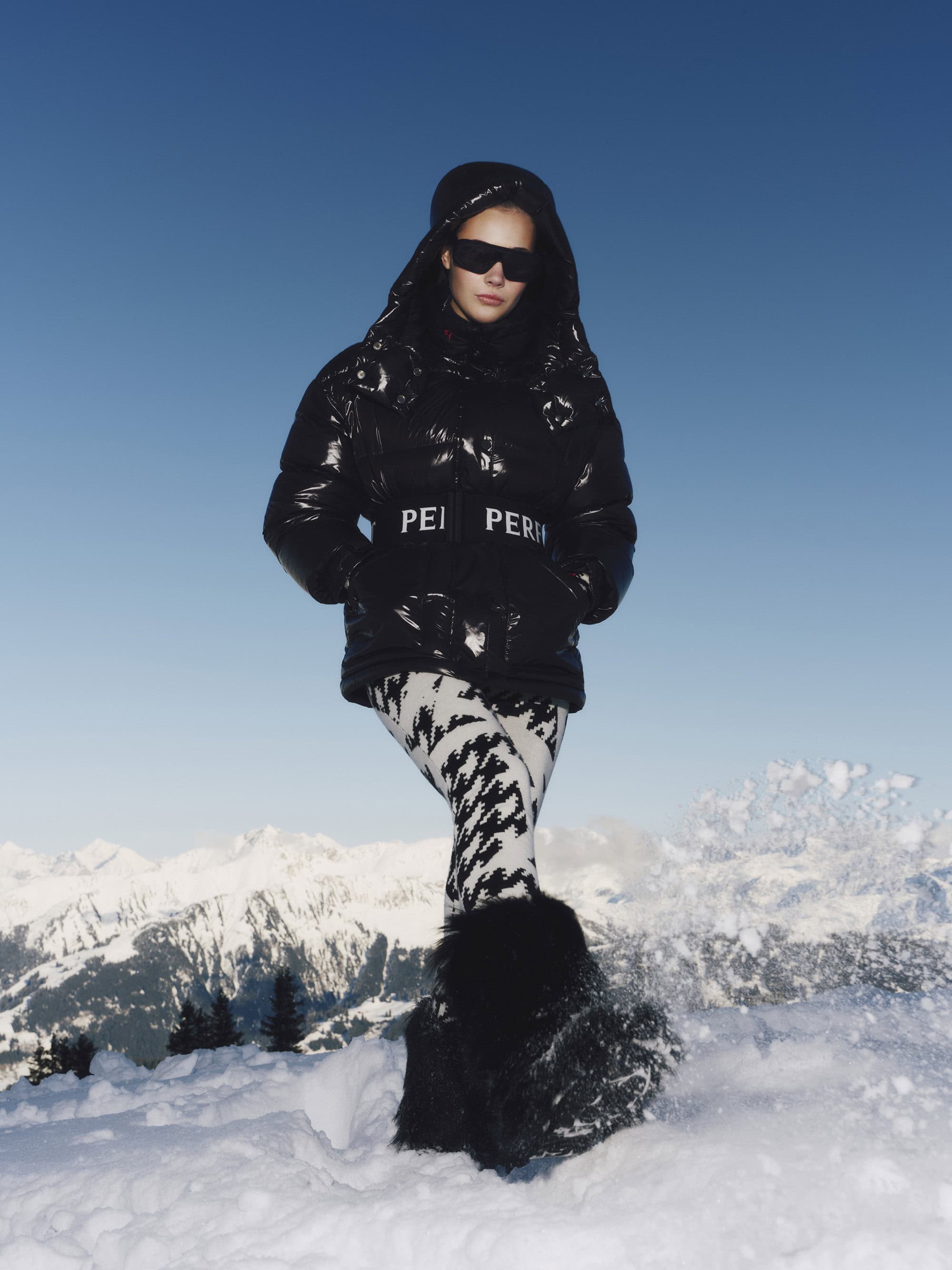 Model wearing a black puffer jacket and patterned pants standing in snow against a mountain backdrop