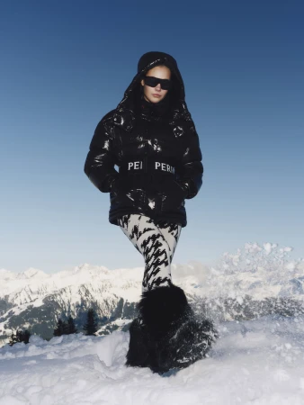 Model wearing a black puffer jacket and patterned pants standing in snow against a mountain backdrop