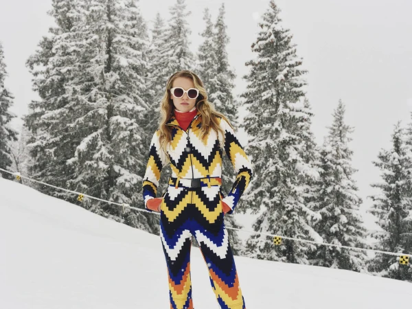 Model wearing a patterned ski suit and white sunglasses standing in a snowy forest for Perfect Moment