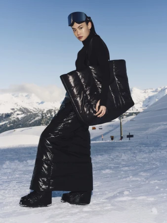 Model in black ski wear holding a large puffy black tote bag against a snowy mountain landscape