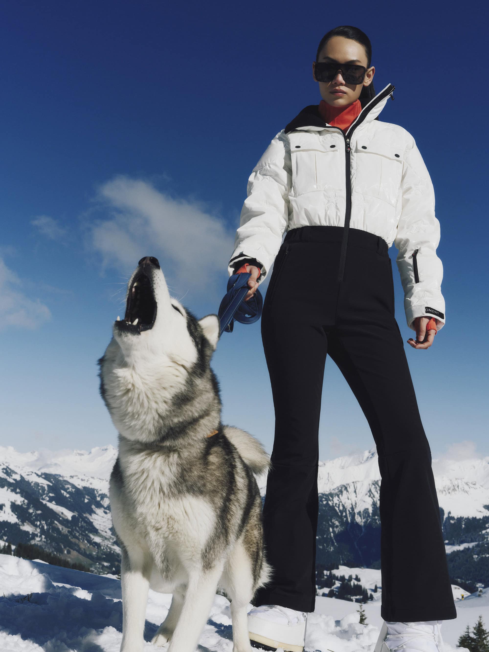 Model in white puffer jacket and black ski pants holding a husky howling outdoors on snow under a clear blue sky
