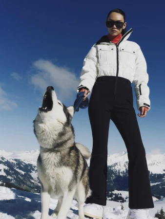 Model in white puffer jacket and black ski pants holding a husky howling outdoors on snow under a clear blue sky