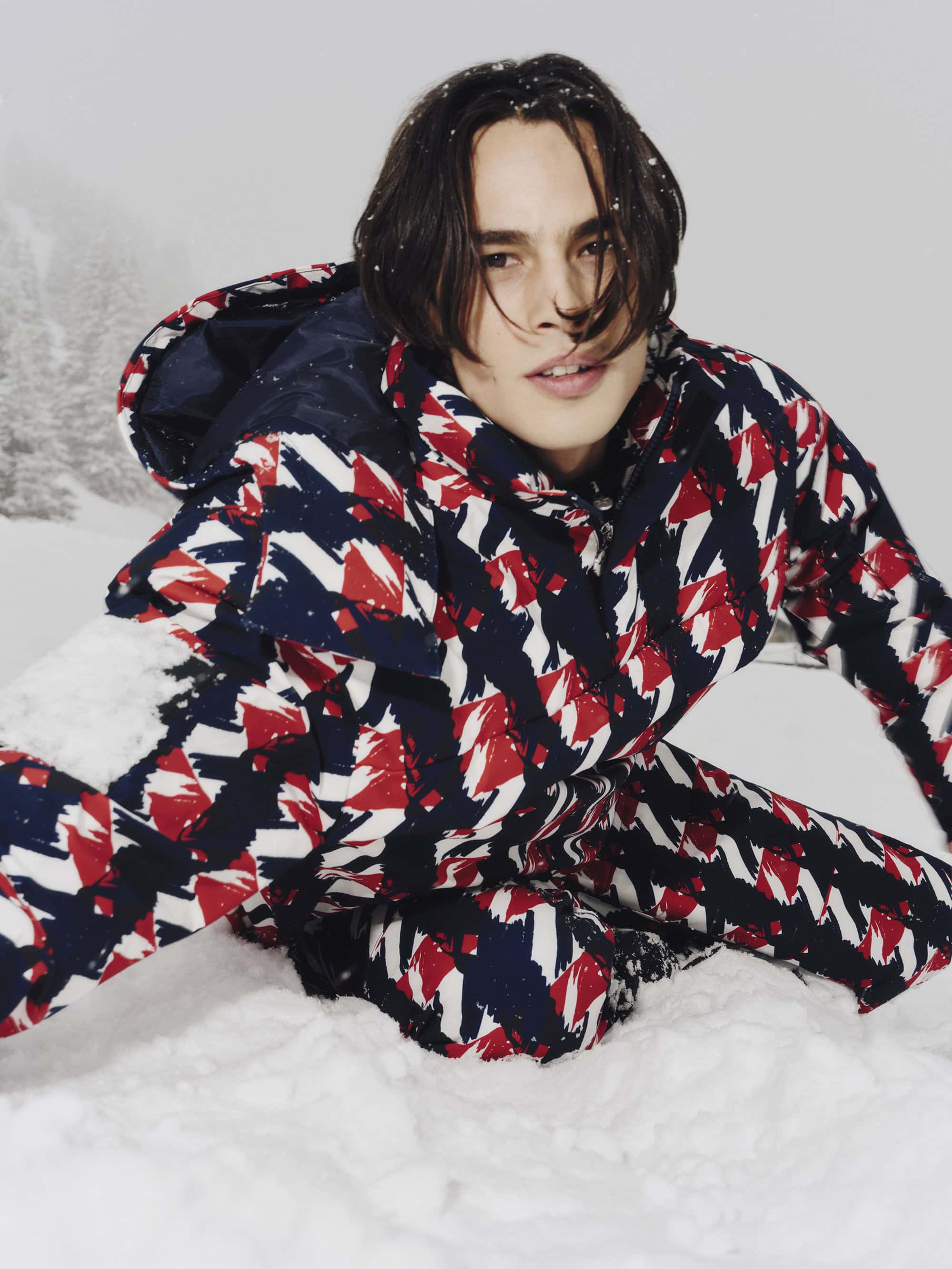 Model wearing a red, white, and blue patterned ski suit kneeling in snow against a snowy mountain backdrop