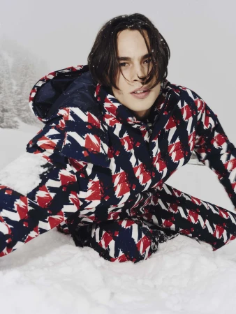 Model wearing a red, white, and blue patterned ski suit kneeling in snow against a snowy mountain backdrop
