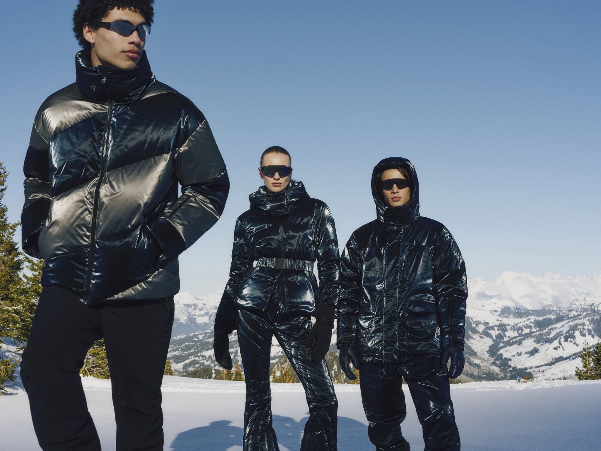 Three models wearing glossy metallic puffer jackets and sunglasses pose outdoors against a snowy mountain landscape under a clear blue sky.