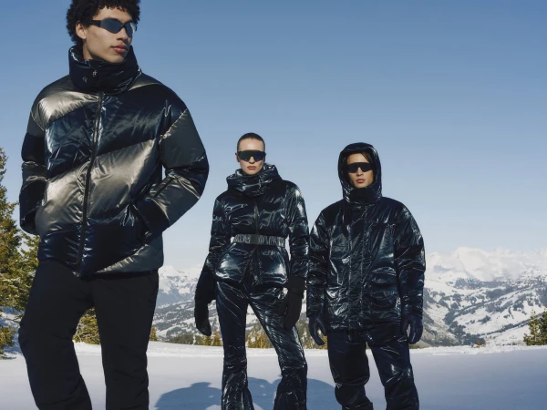 Three models wearing glossy metallic puffer jackets and sunglasses pose outdoors against a snowy mountain landscape under a clear blue sky.