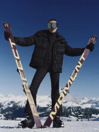 Model in black ski jacket and mirrored goggles holding up two long skis against a snowy mountain backdrop.