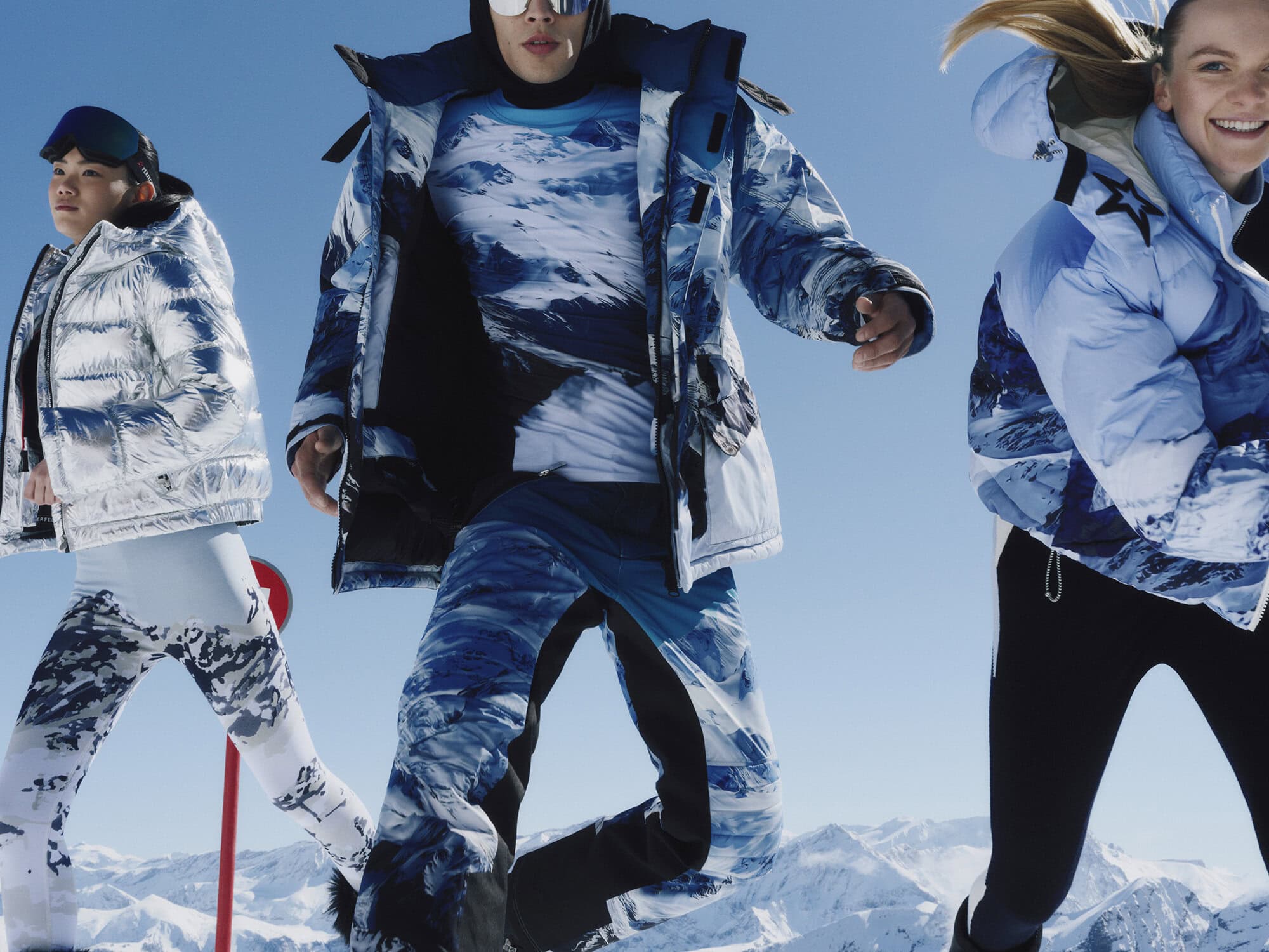 Three models wearing winter ski apparel including blue and silver puffer jackets against a snowy mountain backdrop.