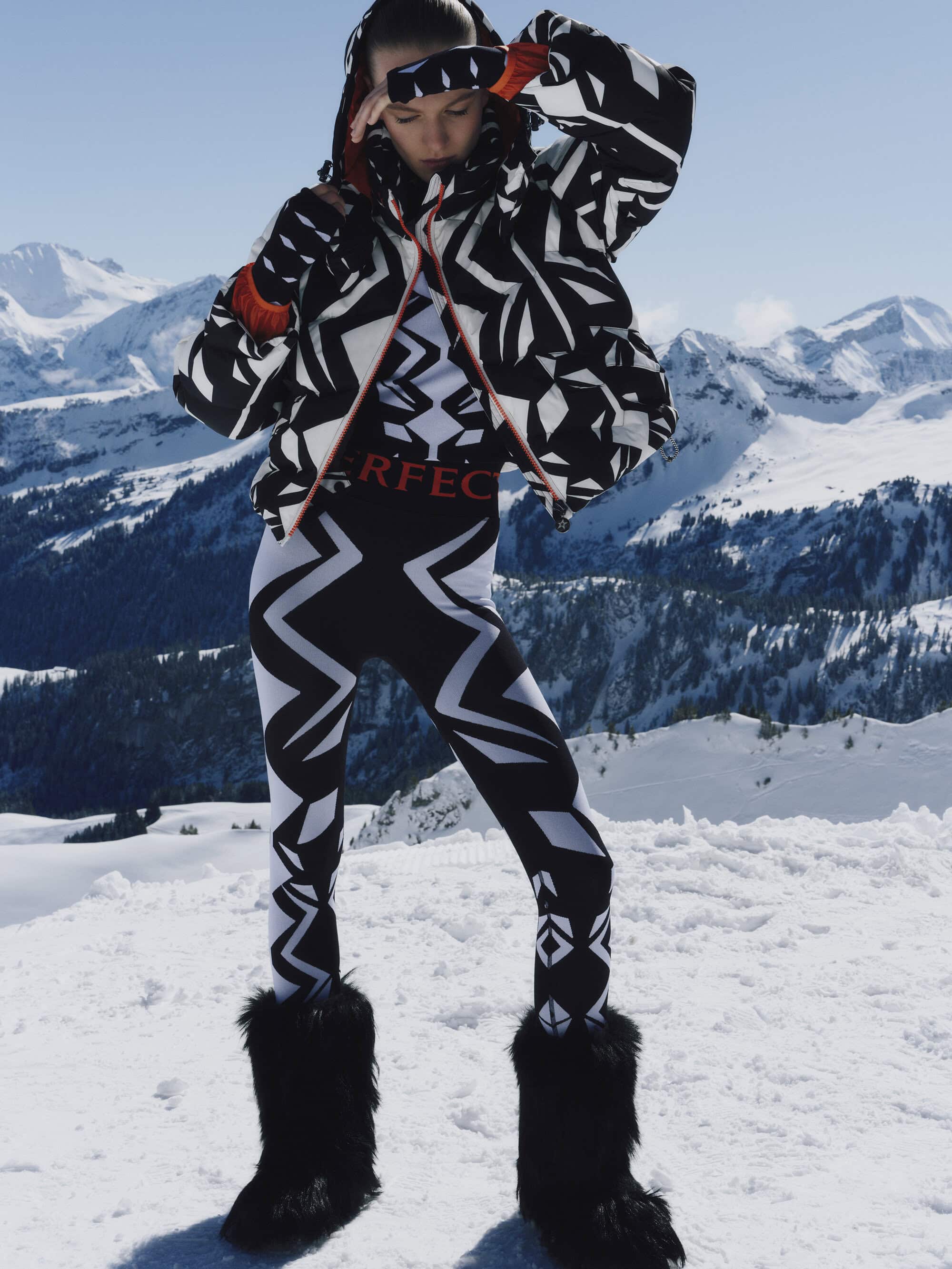 Model wearing black and white geometric ski jacket and leggings with furry boots standing on a snowfield against mountain peaks