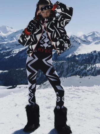 Model wearing black and white geometric ski jacket and leggings with furry boots standing on a snowfield against mountain peaks