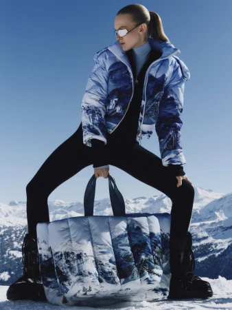 Model in blue mountain-print puffer jacket and matching tote bag standing against snowy peaks under a clear blue sky