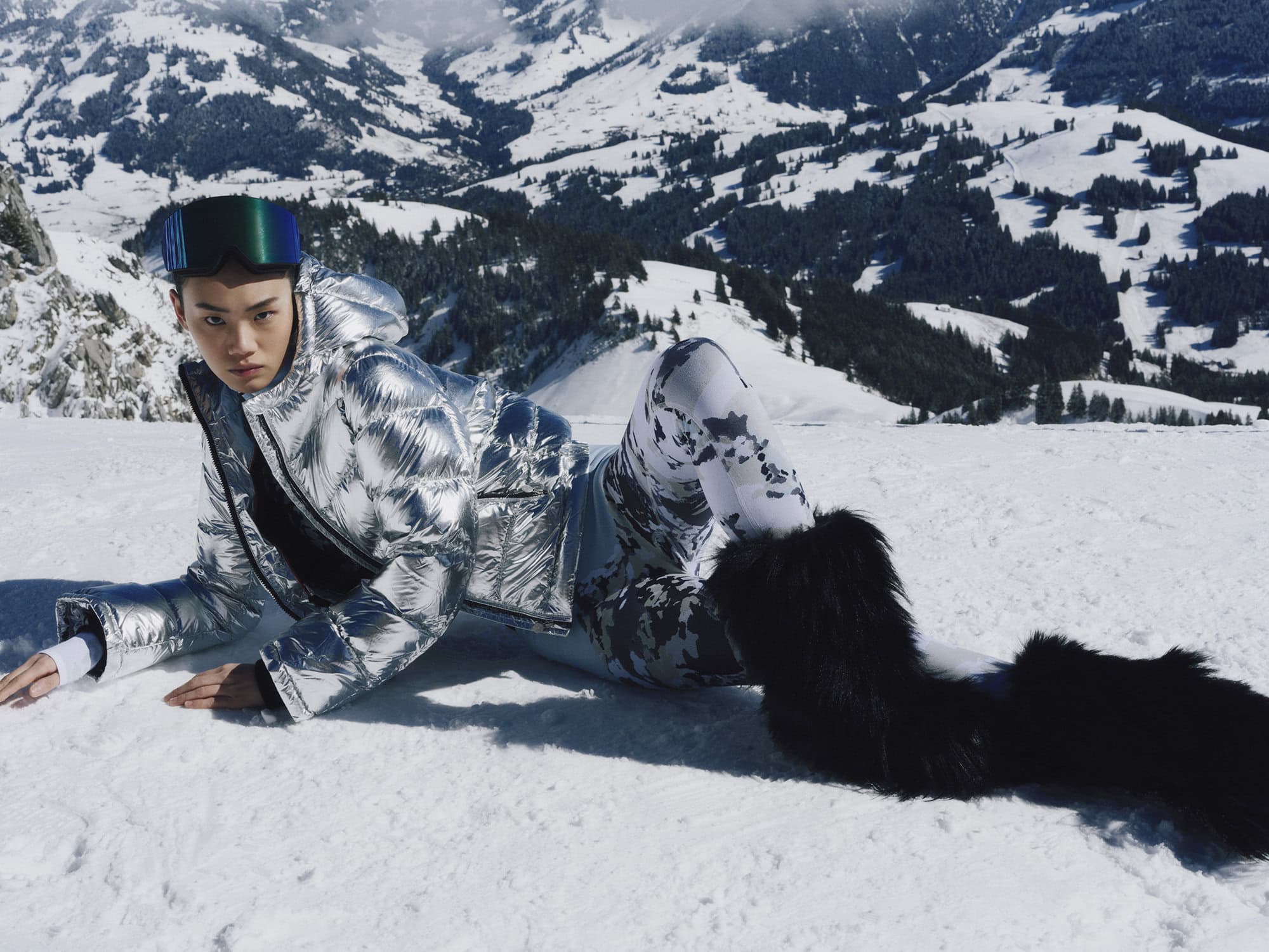 Model wearing a metallic silver puffer jacket and ski goggles lying on a snowy slope in Gstaad