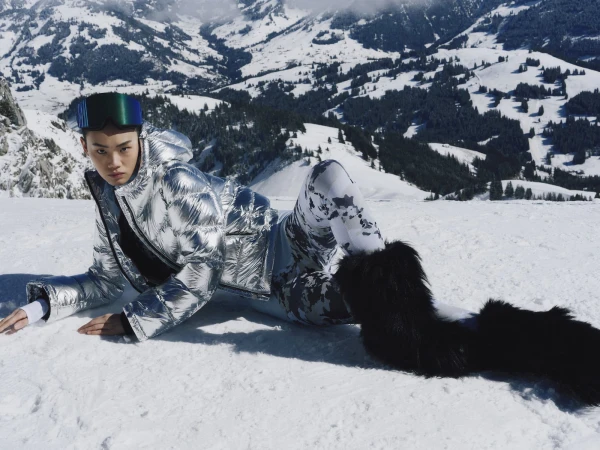 Model wearing a metallic silver puffer jacket and ski goggles lying on a snowy slope in Gstaad