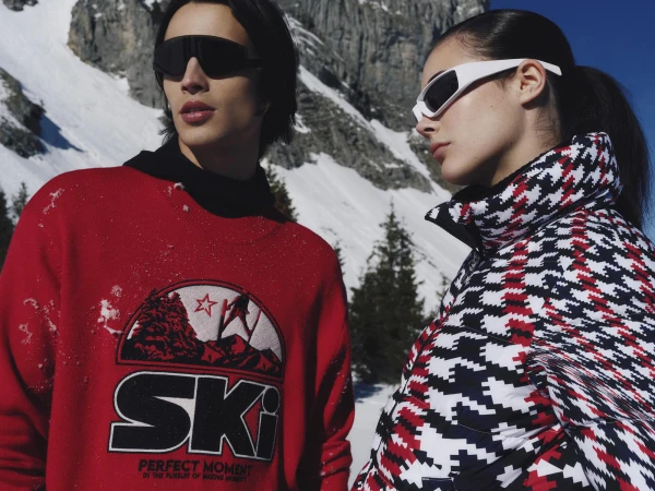 Two models wearing ski apparel and sunglasses outdoors against a snowy mountain background