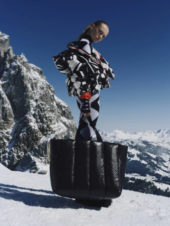 Model wearing black and white patterned winter apparel standing on snowy mountain peaks holding a large black puffy bag