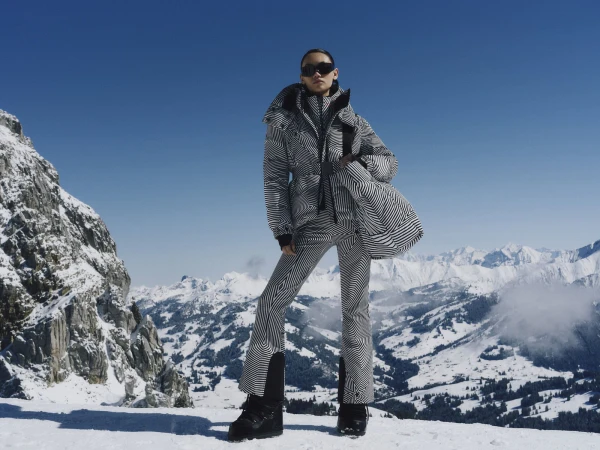 Model wearing a black and white patterned ski suit and sunglasses holding a matching bag against a snowy mountain backdrop.