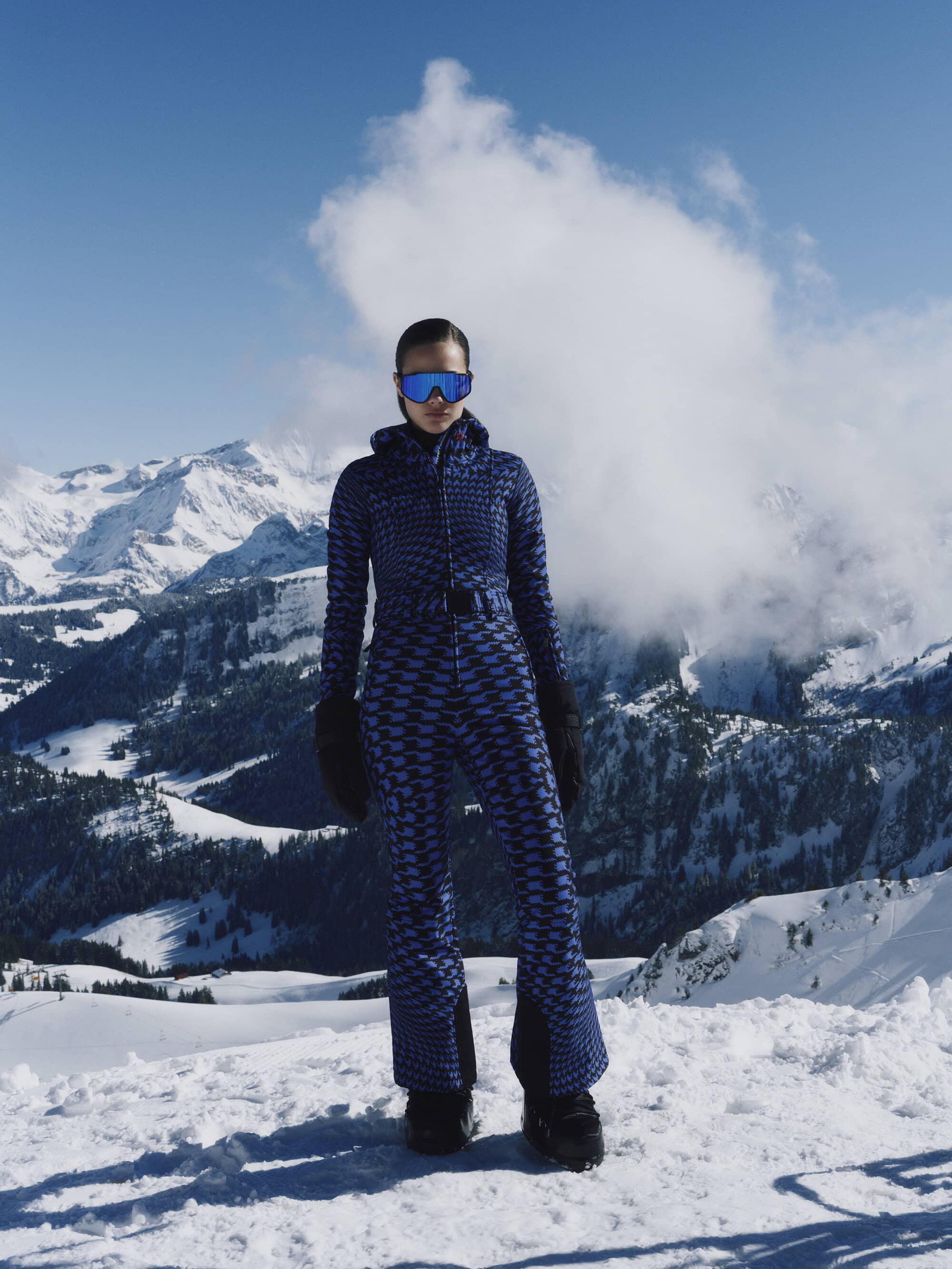 Model wearing a blue patterned ski suit and mirrored goggles standing in snow with mountain peaks behind