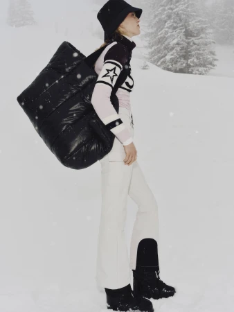 Model wearing white ski pants, patterned sweater, and black bucket hat, carrying a large black puffer bag in a snowy alpine setting