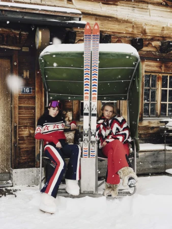 Two female models in winter sportswear seated on an outdoor ski lift bench against a rustic wooden chalet in Gstaad