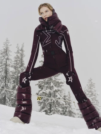 Model wearing a burgundy quilted ski suit with star motifs and puffy boots standing on a snowy slope
