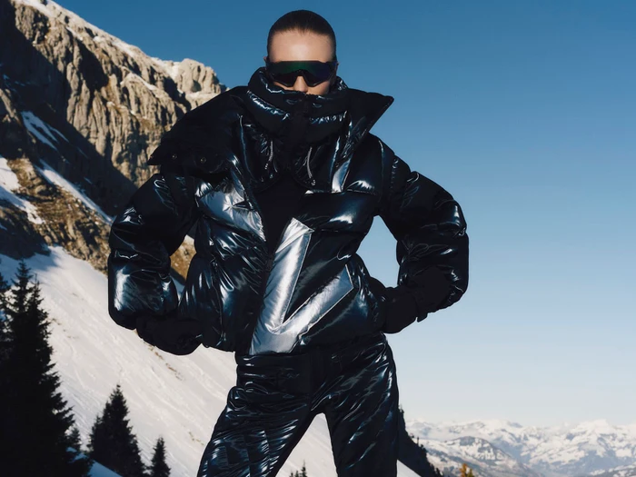 Model wearing a shiny puffer jacket and sunglasses against a snowy mountain and clear blue sky backdrop