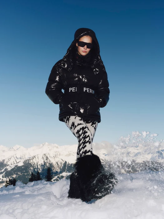 Model wearing a black puffer jacket and patterned pants standing in snow against a mountain backdrop