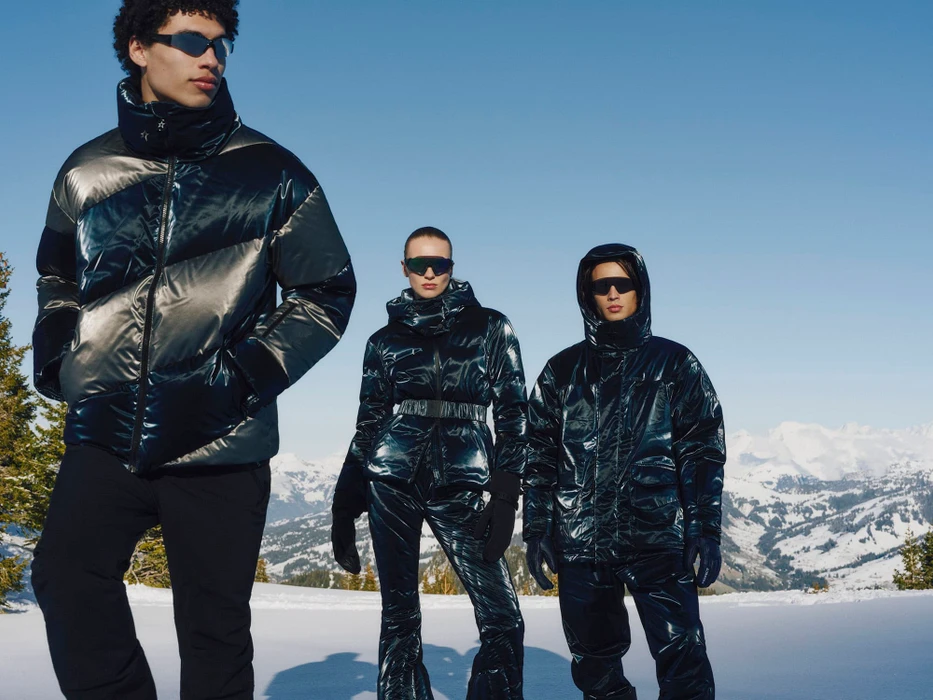 Three models wearing glossy metallic puffer jackets and sunglasses pose outdoors against a snowy mountain landscape under a clear blue sky.