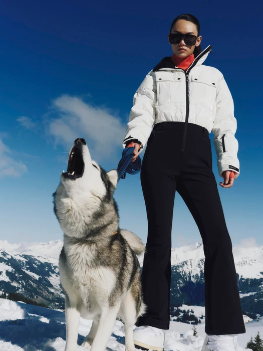 Model in white puffer jacket and black ski pants holding a husky howling outdoors on snow under a clear blue sky