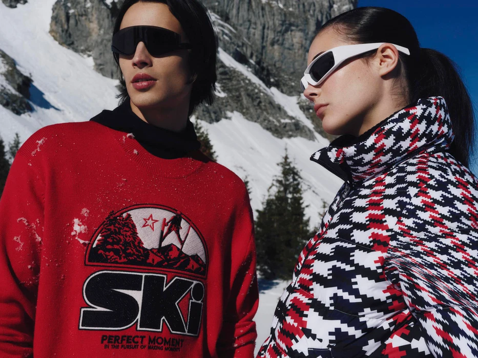 Two models wearing ski apparel and sunglasses outdoors against a snowy mountain background