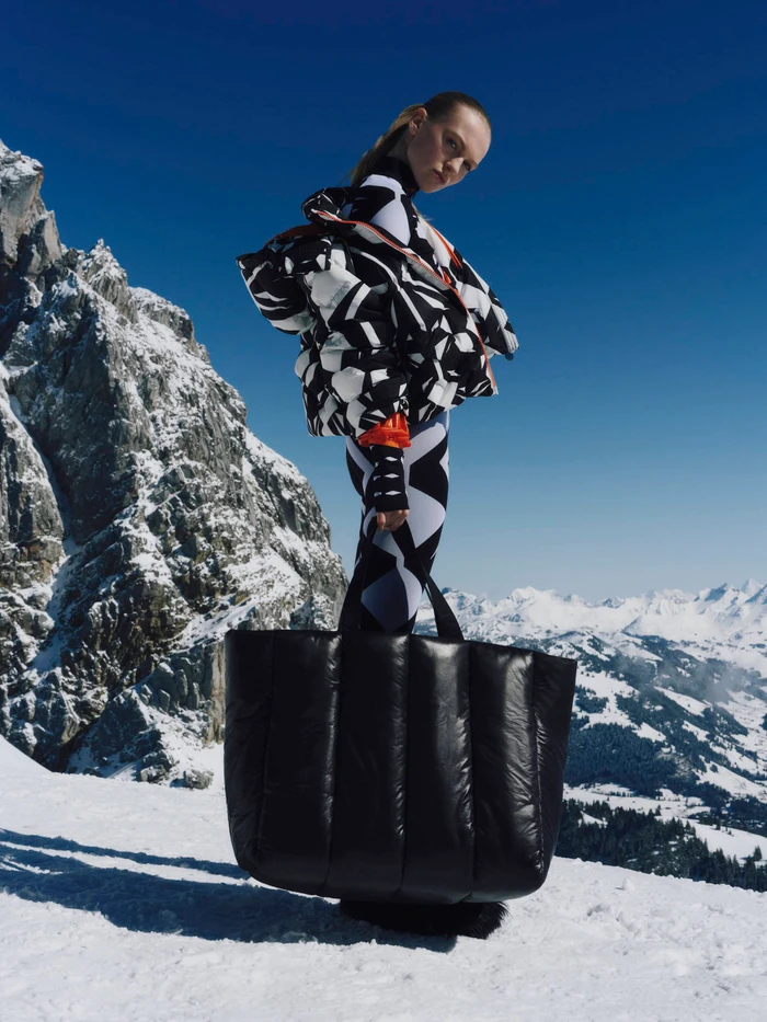 Model wearing black and white patterned winter apparel standing on snowy mountain peaks holding a large black puffy bag