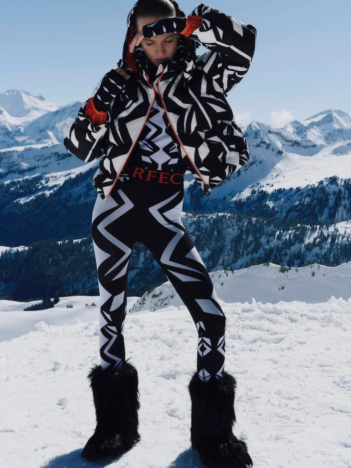 Model wearing black and white geometric ski jacket and leggings with furry boots standing on a snowfield against mountain peaks