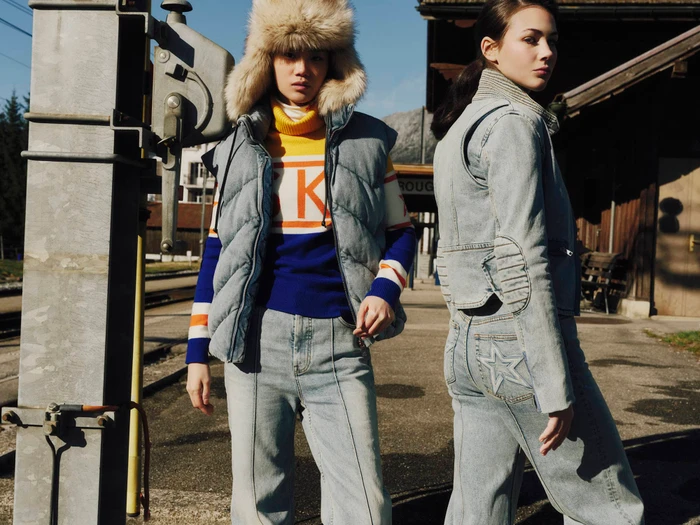 Two models wearing winter puffer vests and denim apparel outdoors near a railway platform for Perfect Moment.
