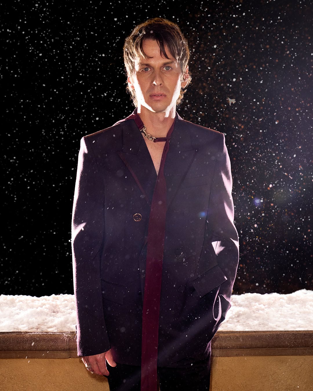 Man wearing a dark plum blazer standing outdoors against a night snow scene for the Gucci Holiday 2024 campaign.