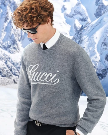 Man with curly brown hair wearing sunglasses and a grey Gucci sweater against a snowy mountain backdrop.