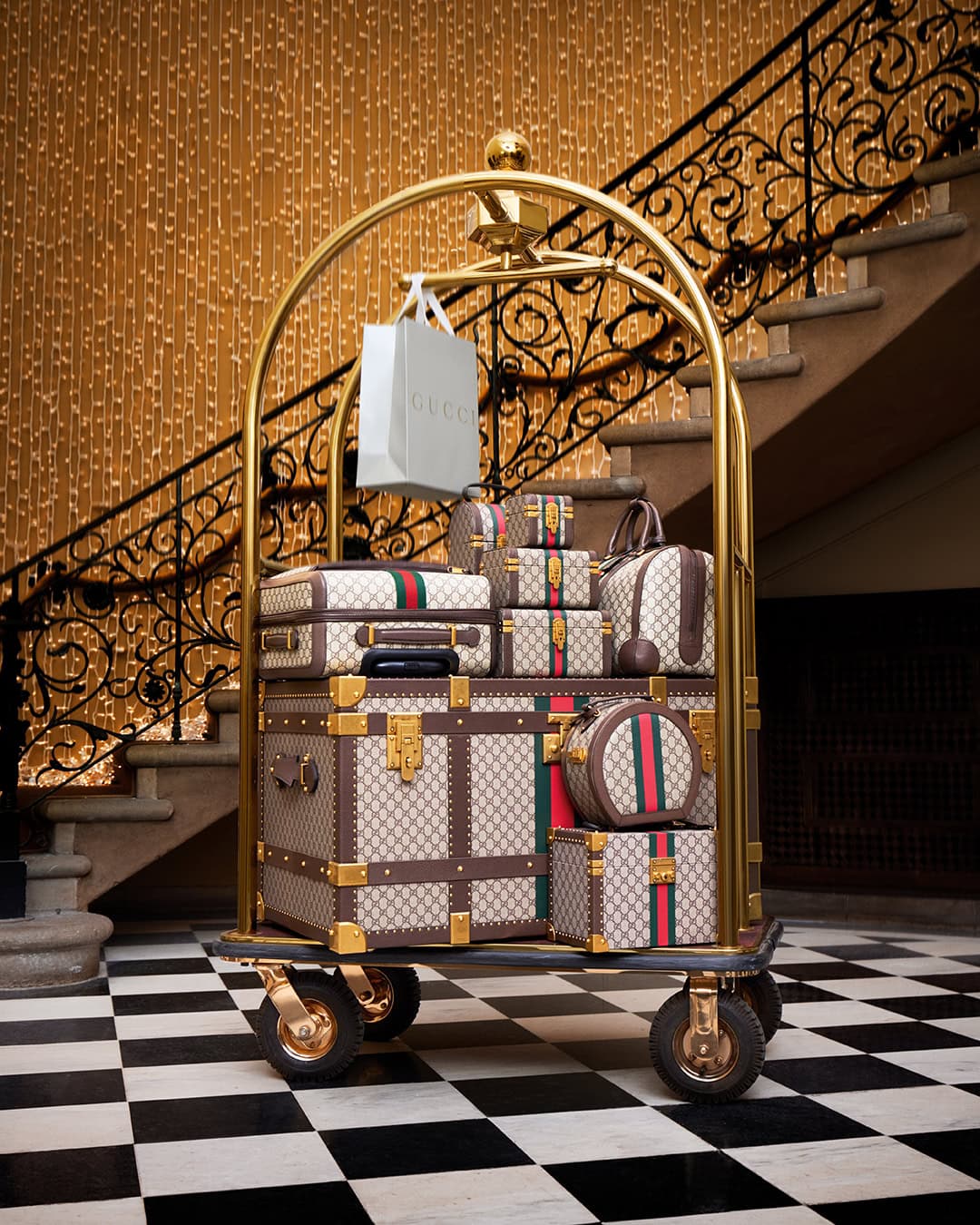 A brass bellhop cart loaded with Gucci monogram luggage and a shopping bag in a grand hotel lobby with a black and white checkerboard floor.