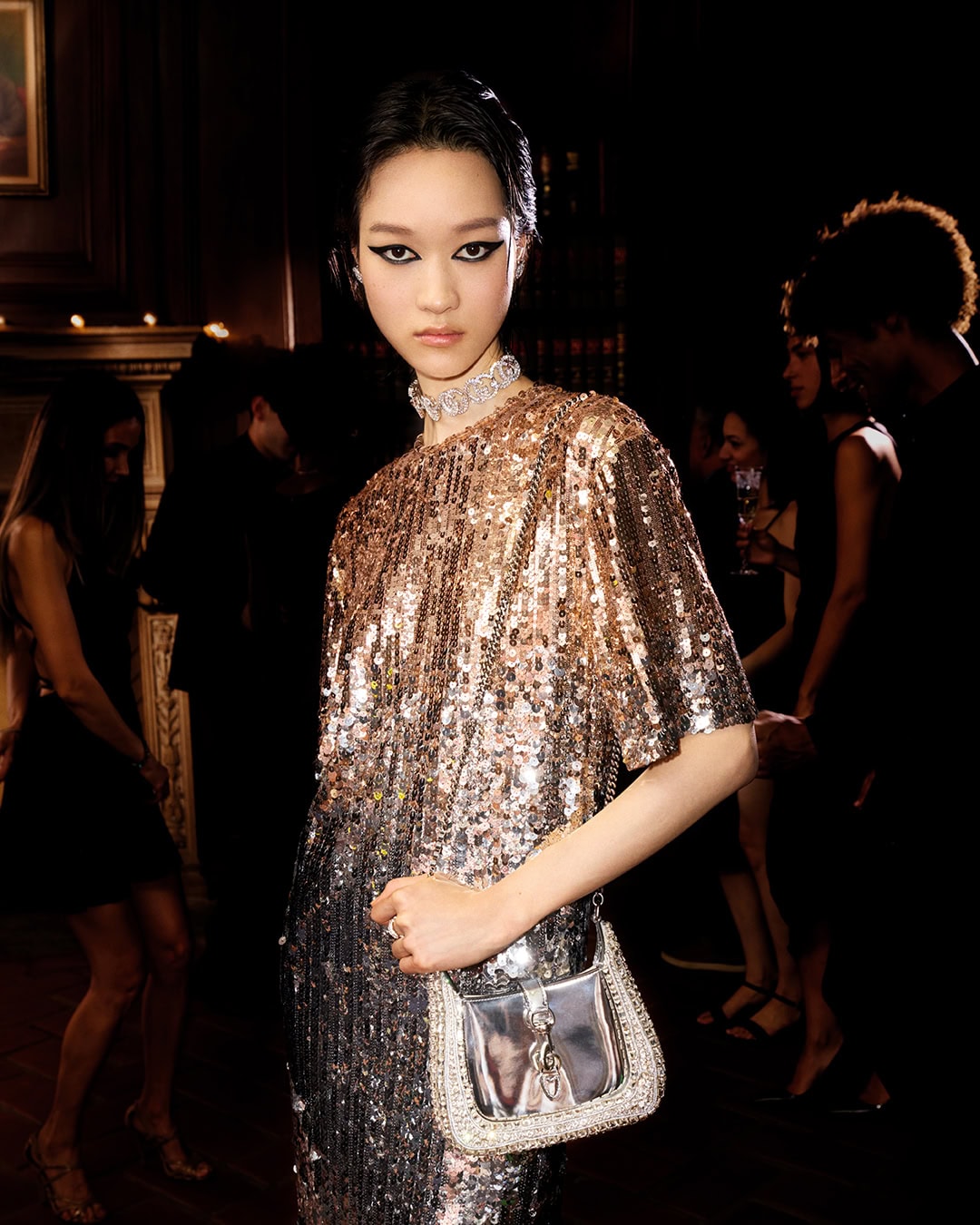 Model in glittering sequin dress wearing diamond choker holding a silver handbag in dim interior