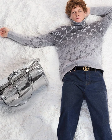 Model in grey Gucci monogram turtleneck and dark denim jeans lying on white snow with a silver duffel bag.