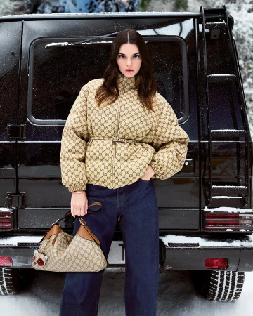 Kendall Jenner wearing a monogram puffer jacket and holding a handbag in front of a black vehicle in the snow.