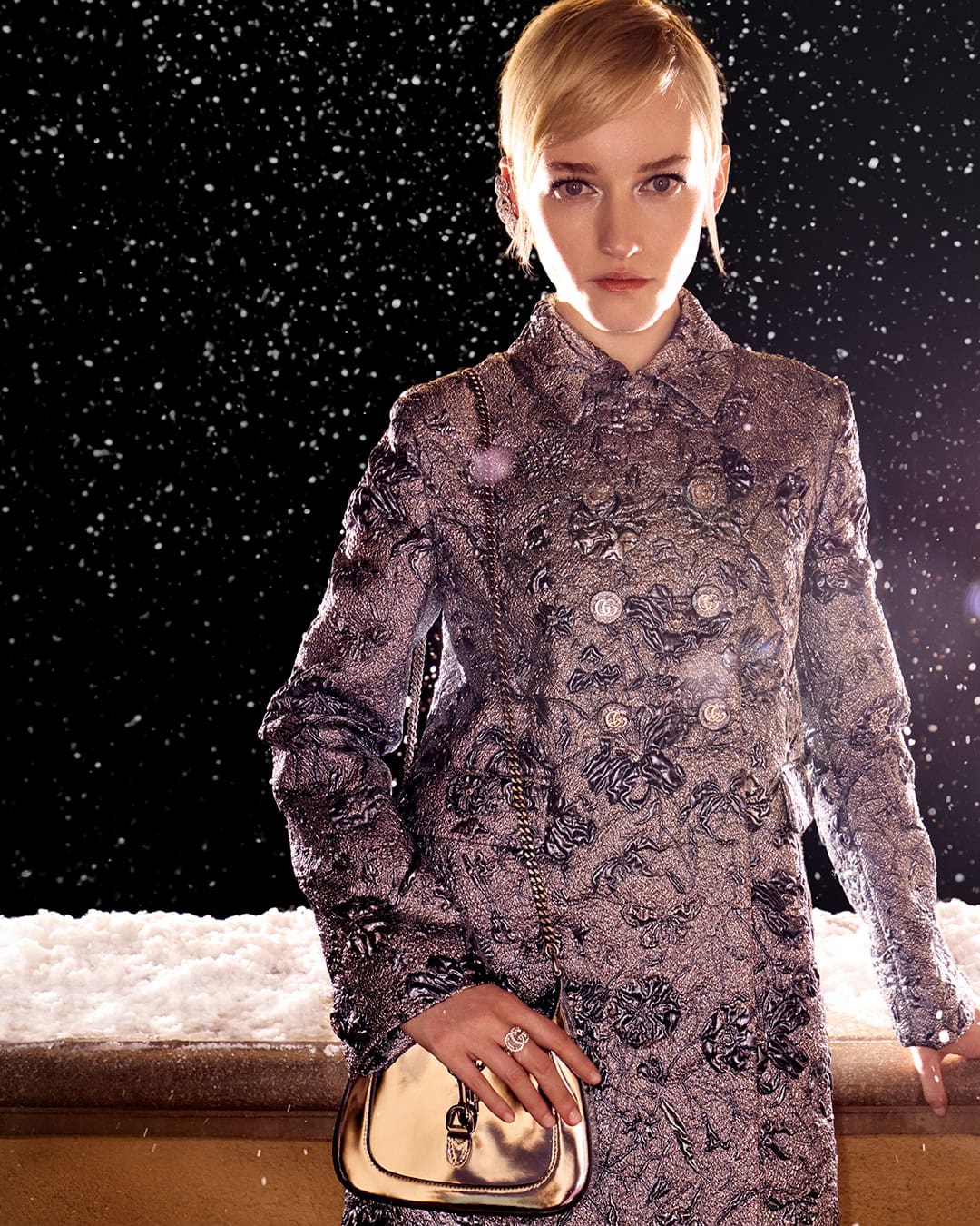 Model in patterned grey double breasted coat holding metallic gold shoulder bag outdoors at night in snow