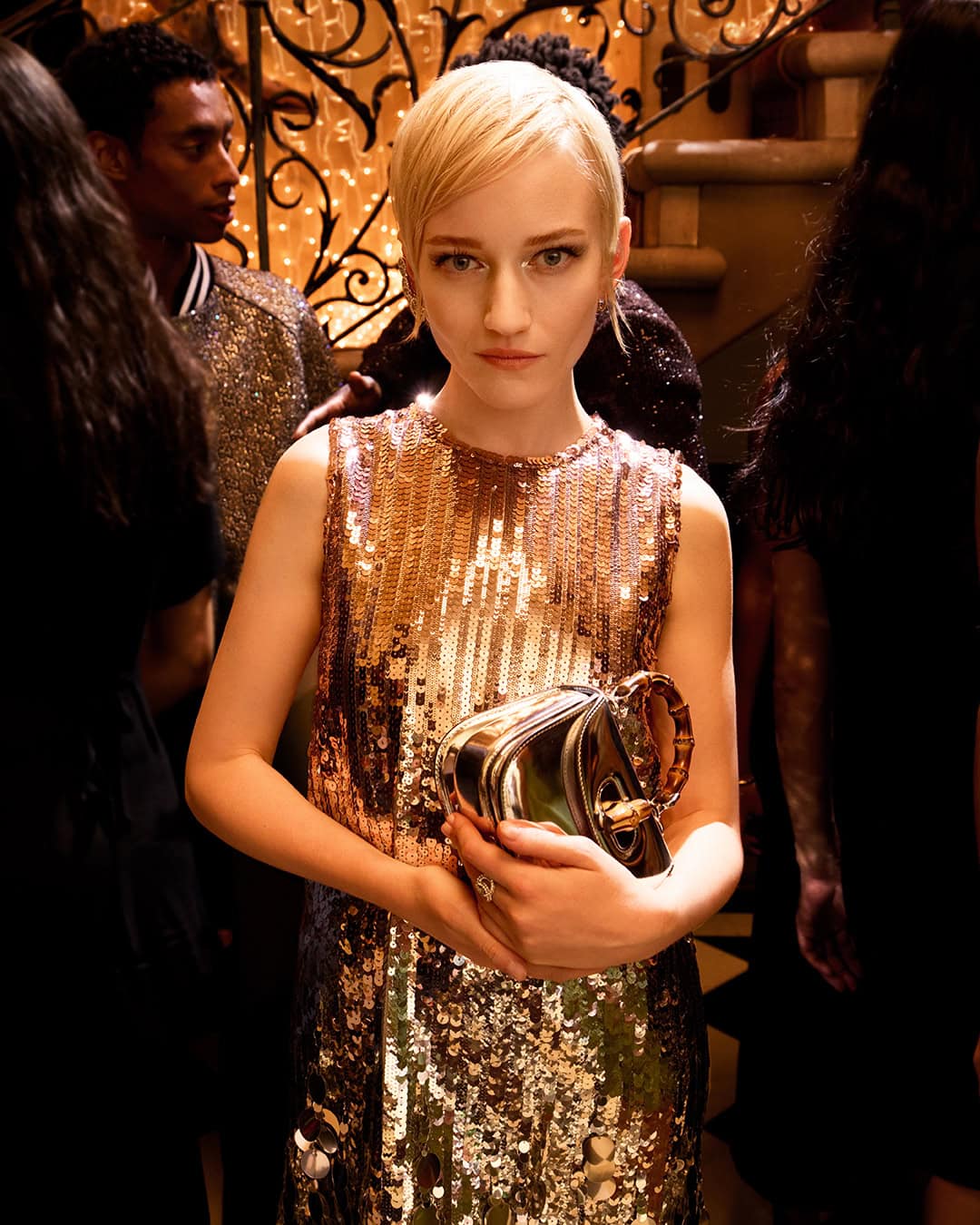 Blonde woman wearing a shimmering sequin dress holding a metallic handbag in a festive interior setting for Gucci Holiday Campaign.