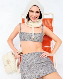 Kendall Jenner wearing a patterned monogram crop top and skirt set while seated on an orange folding chair against a white background.