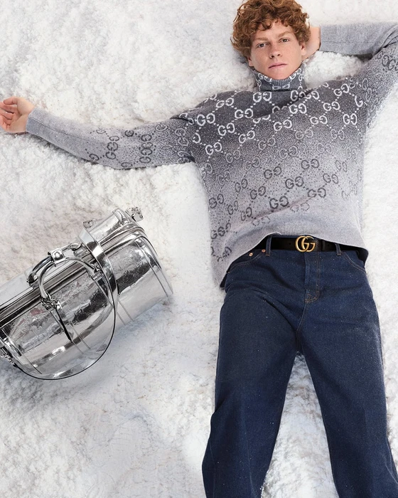 Model in grey Gucci monogram turtleneck and dark denim jeans lying on white snow with a silver duffel bag.