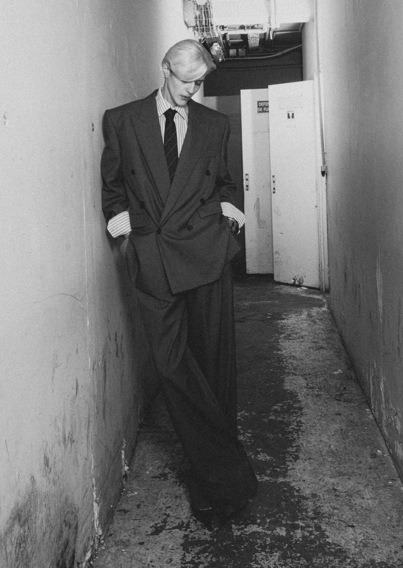 A monochrome fashion shot of a young adult male leaning against a wall in a wide, double-breasted suit in a narrow hallway with doors.