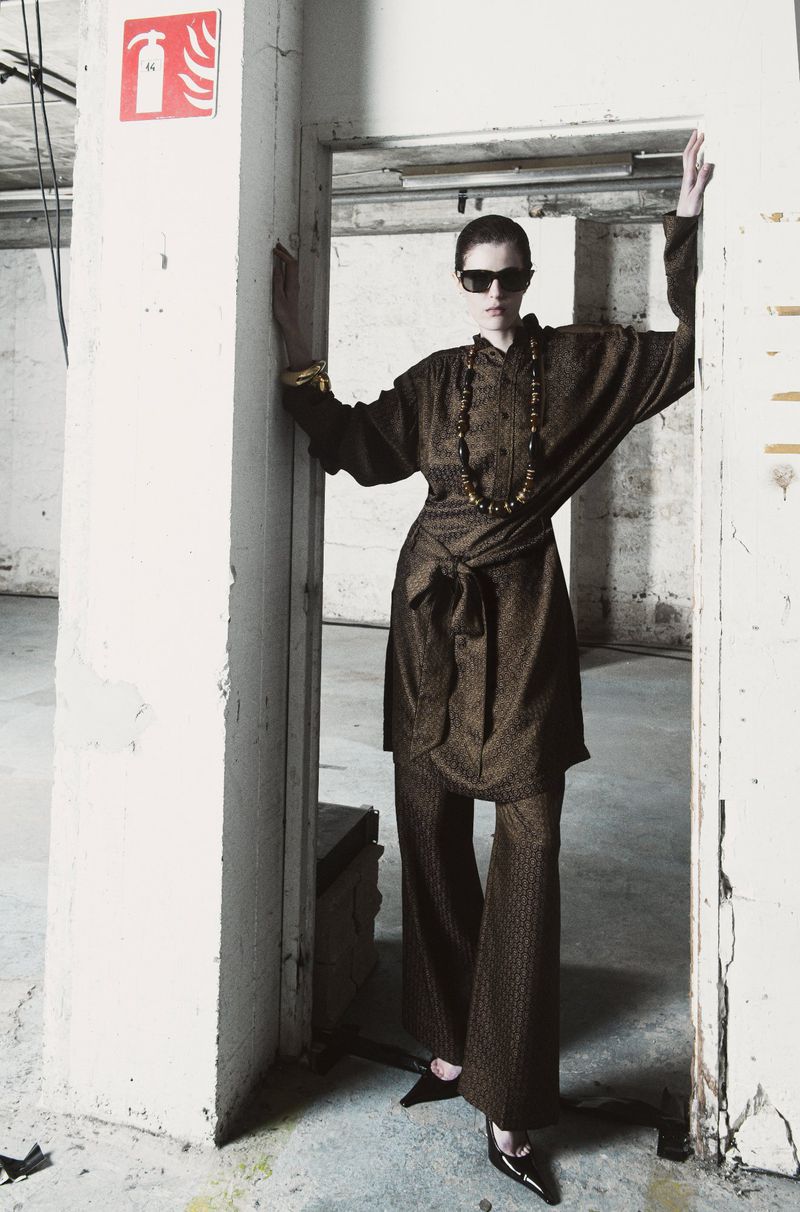 Woman in brown pantsuit with long sleeves, statement necklace, and sunglasses poses in a doorway with arms outstretched, fashion shot.