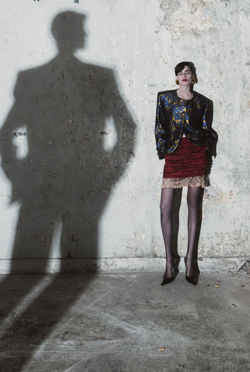 Fashionable woman poses in a jacket and skirt against a textured wall with a shadow, creating an editorial and artistic atmosphere.