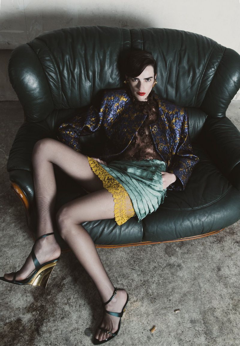 A woman in a blue patterned jacket sits on a green leather chair, wearing a pleated skirt, hosiery, and sandals in a fashion shot.