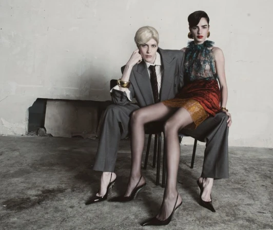 Two women pose in a photo studio. One sits on a chair wearing a gray suit; the other sits on her lap wearing a short red dress.