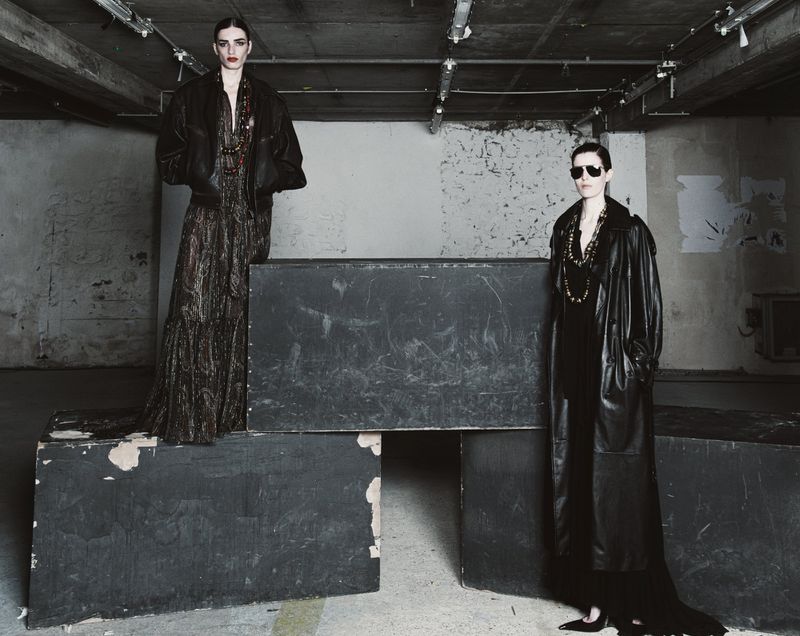 Two women in long coats and necklaces pose in a gritty urban setting, illuminated by overhead lighting, showcasing dark, elegant fashion.