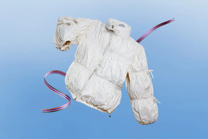 White puffer jacket with gathered sleeves floating against a gradient blue background with curving striped ribbons