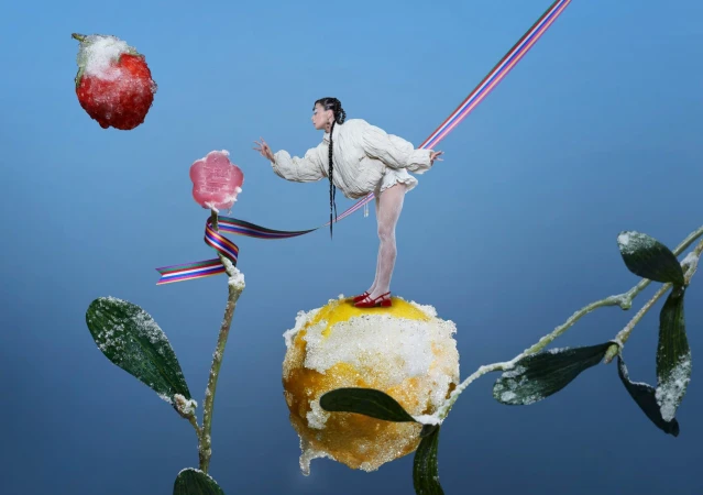 Model standing on a frosted citrus fruit with floating strawberry and candy against a blue background for Sundae School Holiday 2025.